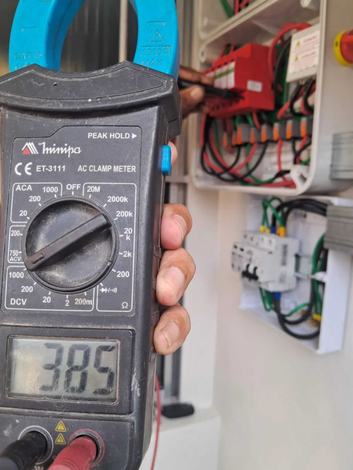 Alicate amperímetro em primeiro plano, com componentes elétricos ao fundo.